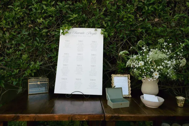 Table with wedding seating chart, framed sign, and white flowers against a green hedge backdrop