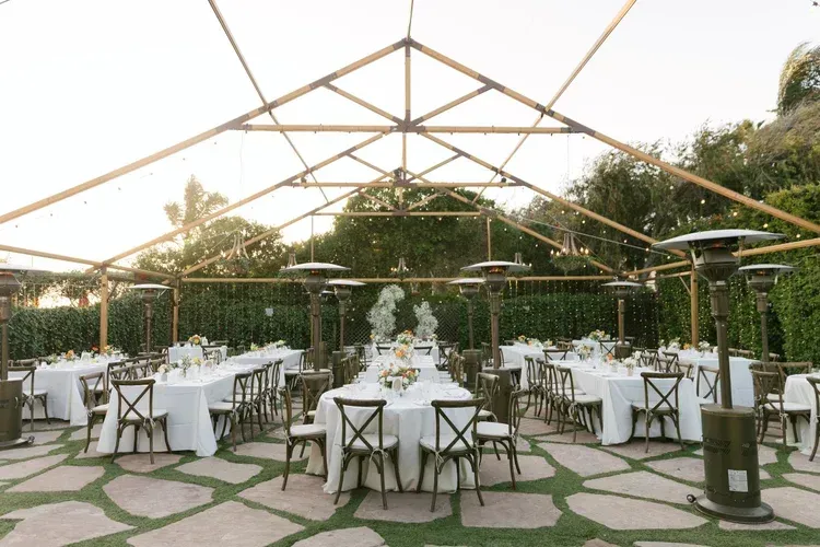 Outdoor banquet setup with white tables, chairs, and a canopy under string lights in a garden setting