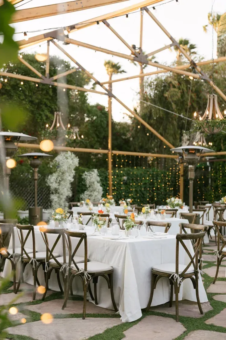 Outdoor wedding reception with white-clothed tables, wooden chairs, and string lights in a garden pavilion