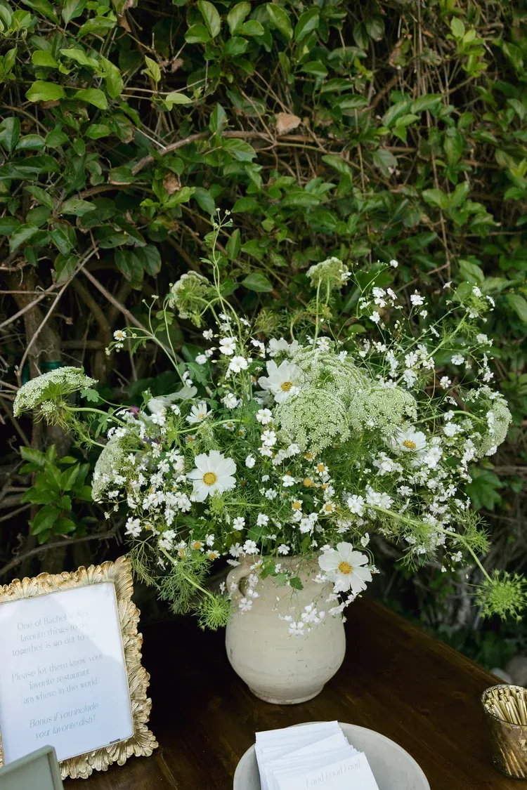 White vase of small white flowers on a wooden table against a leafy green wall