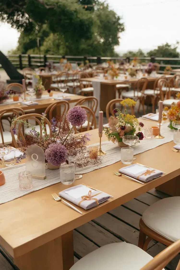 Elegant outdoor dining table set with white plates, purple flowers, and gold accents under trees