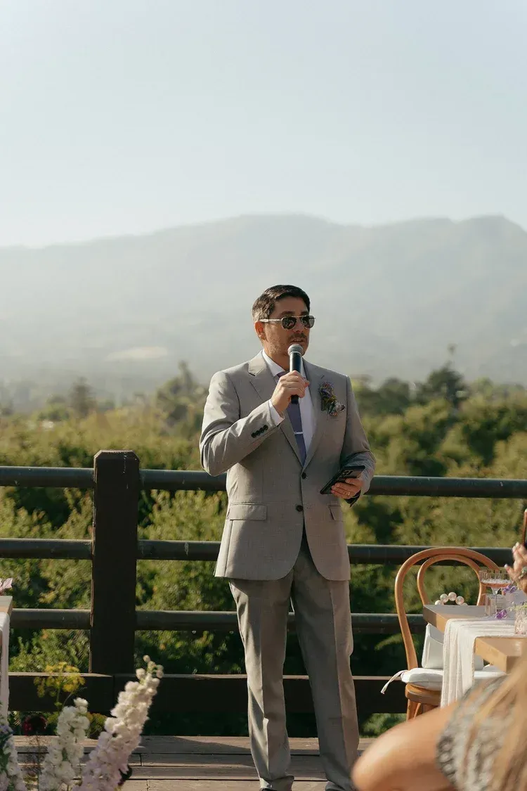 Man in a light gray suit speaking into a microphone outdoors beside a table and railing, with mountains behind him