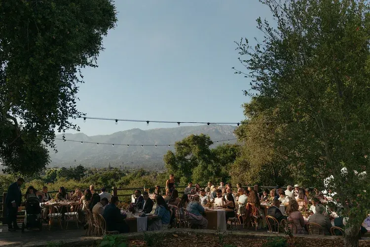 Outdoor garden wedding reception with seated guests, string lights, and trees at sunset.