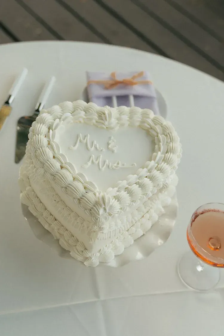 Heart-shaped white frosted cake on a table, with candles and a glass of rose drink nearby.