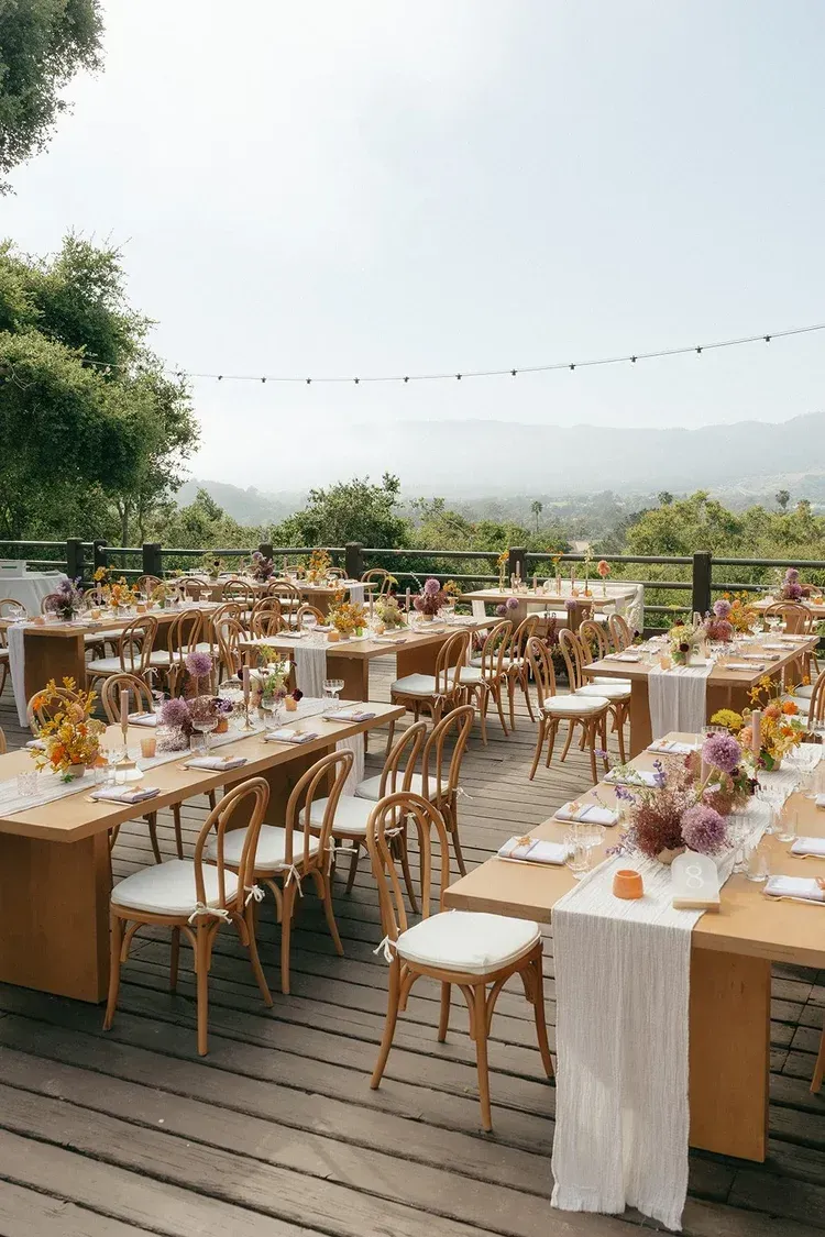 Outdoor banquet tables set on a wooden deck with white table runners, flowers, and string lights