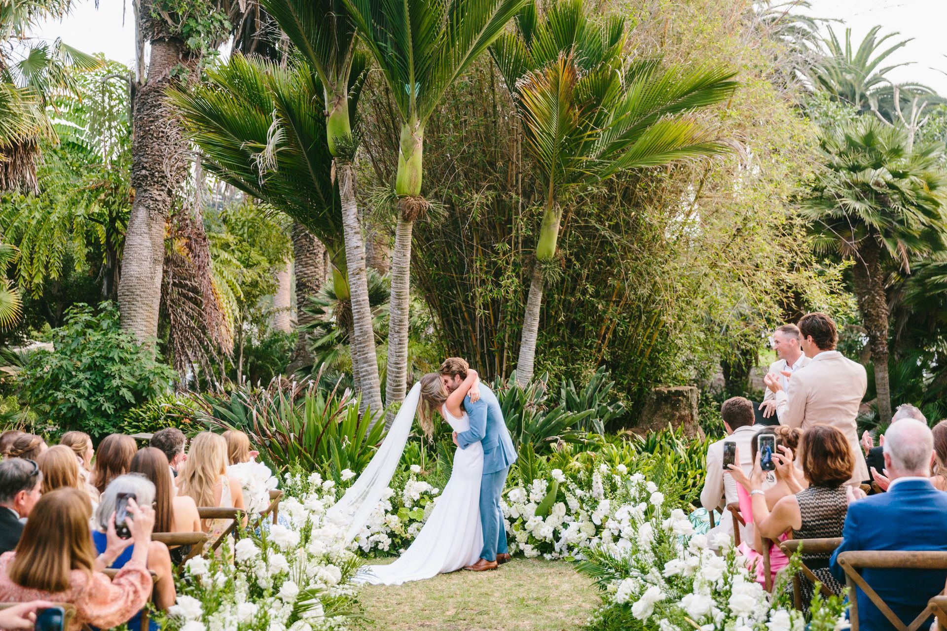 Wedding couple kissing in a lush garden aisle lined with white flowers and seated guests