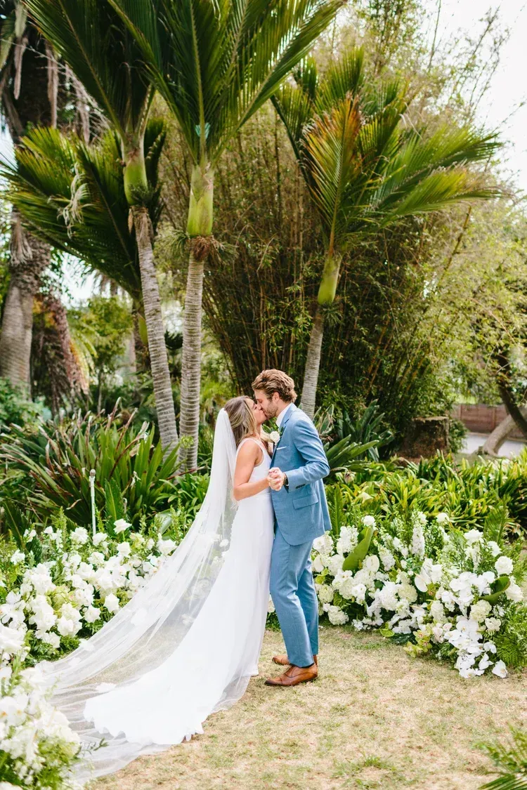 Wedding couple kissing in a lush garden, bride in white gown and groom in blue suit, surrounded by white flowers