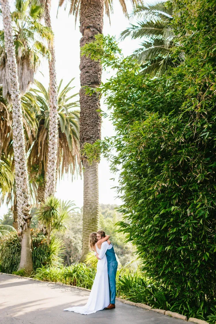 Bride and groom embracing on a palm-lined path beside tall greenery