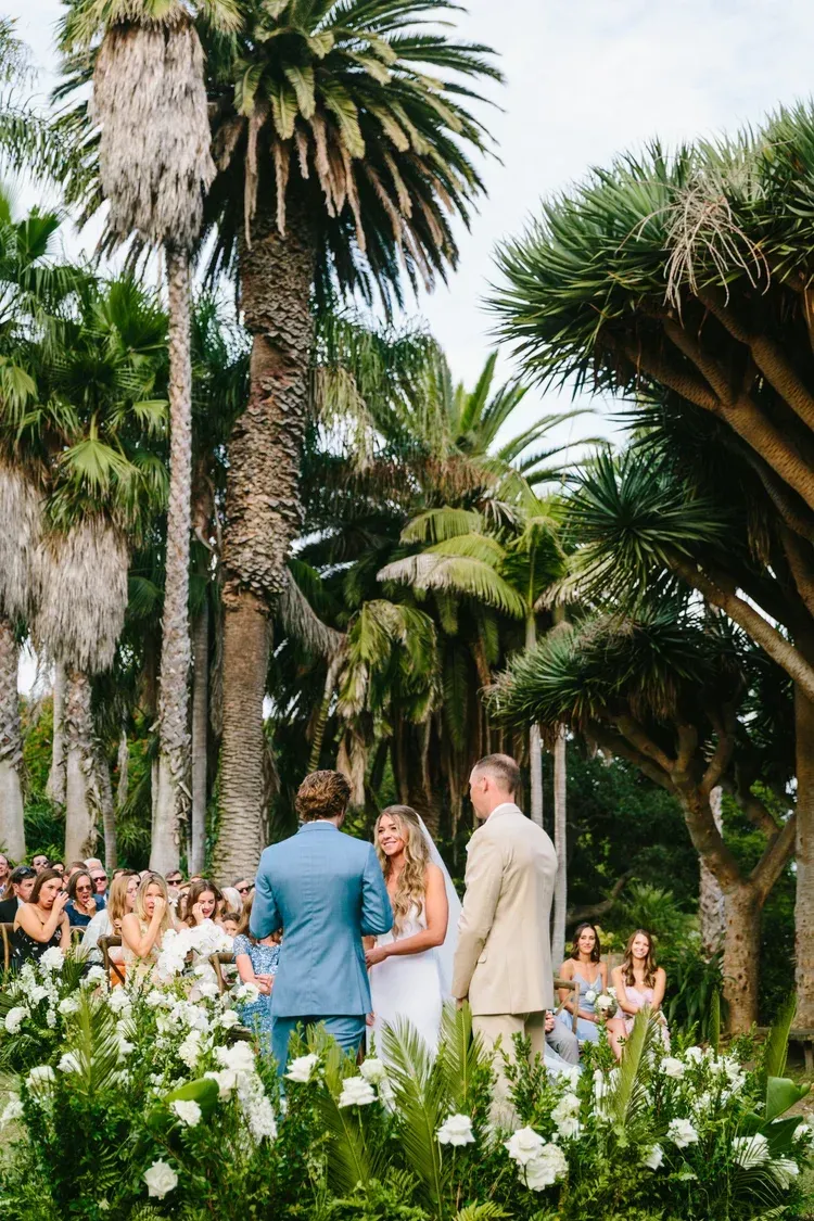 Wedding ceremony in a lush tropical garden, with a couple and officiant among palm trees and white flowers