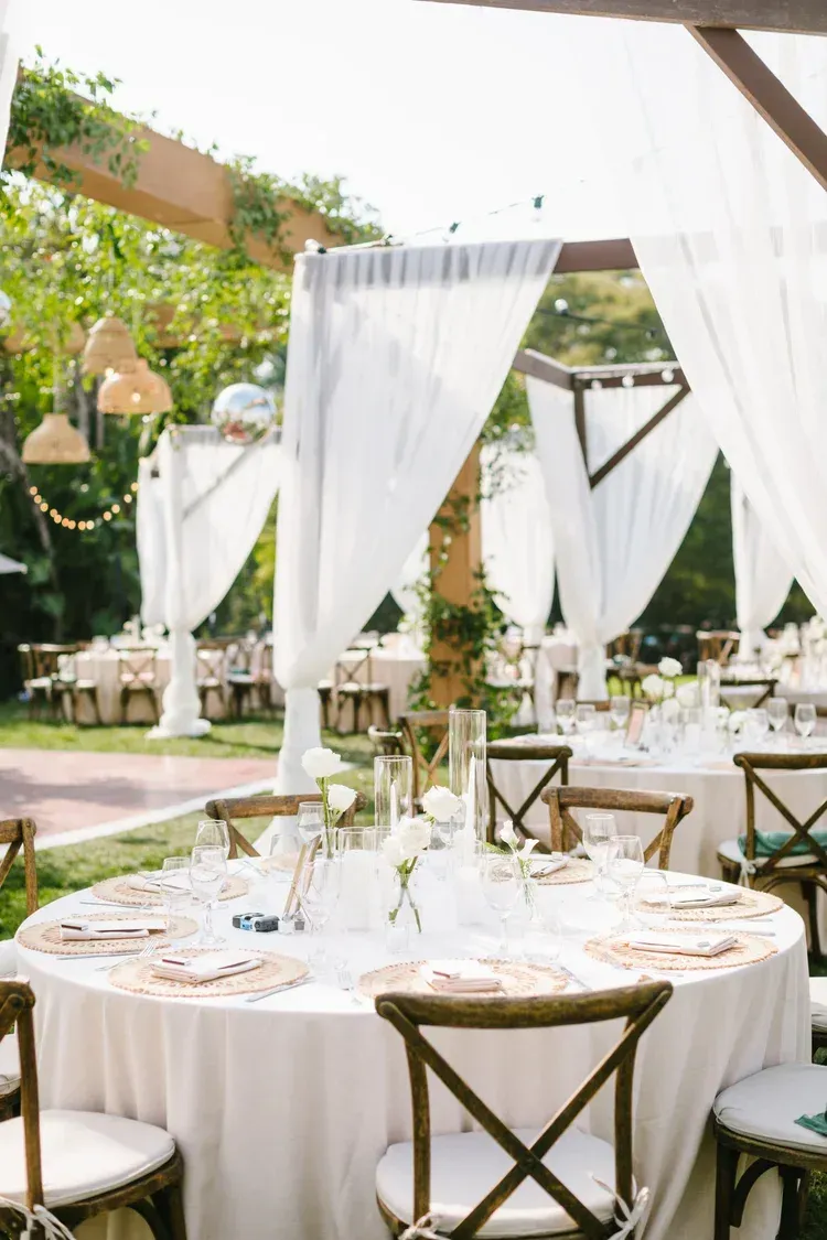 Elegant outdoor wedding reception with white drapes, round tables, and gold-accented chairs under a canopy.