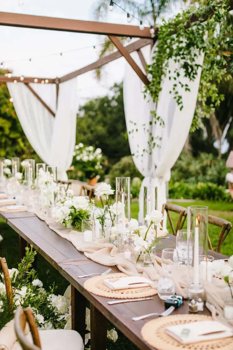 Outdoor wedding reception table with white decor, candles, and greenery under a wooden pergola