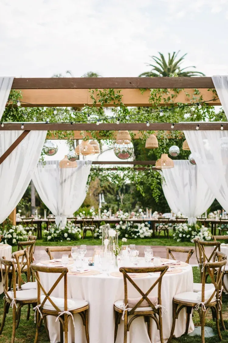 Elegant outdoor wedding reception with round tables, white drapery, and lush greenery under a wooden canopy