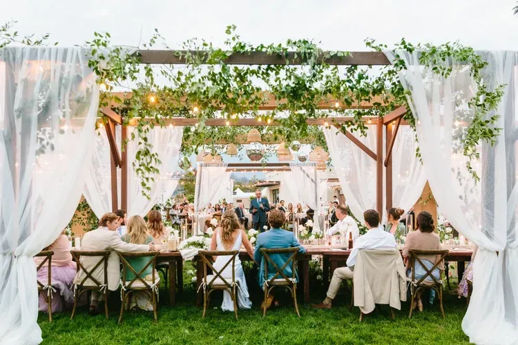 Outdoor garden wedding reception under a floral arbor with guests seated at long tables on the lawn