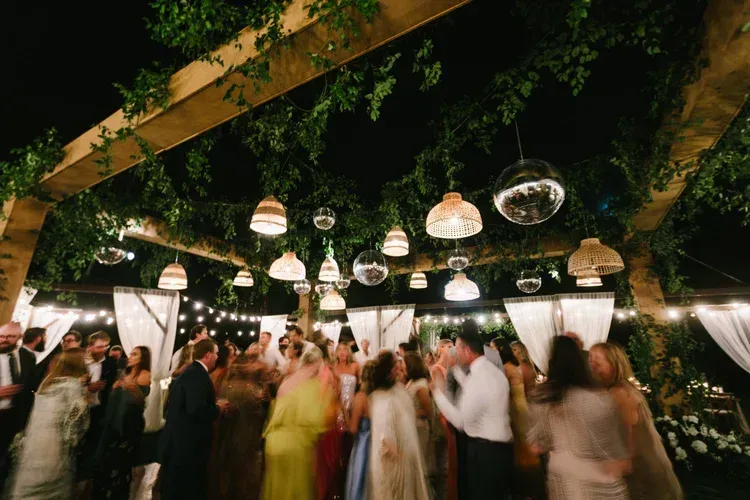 Guests dance under hanging lights and greenery at a warmly lit wedding reception.
