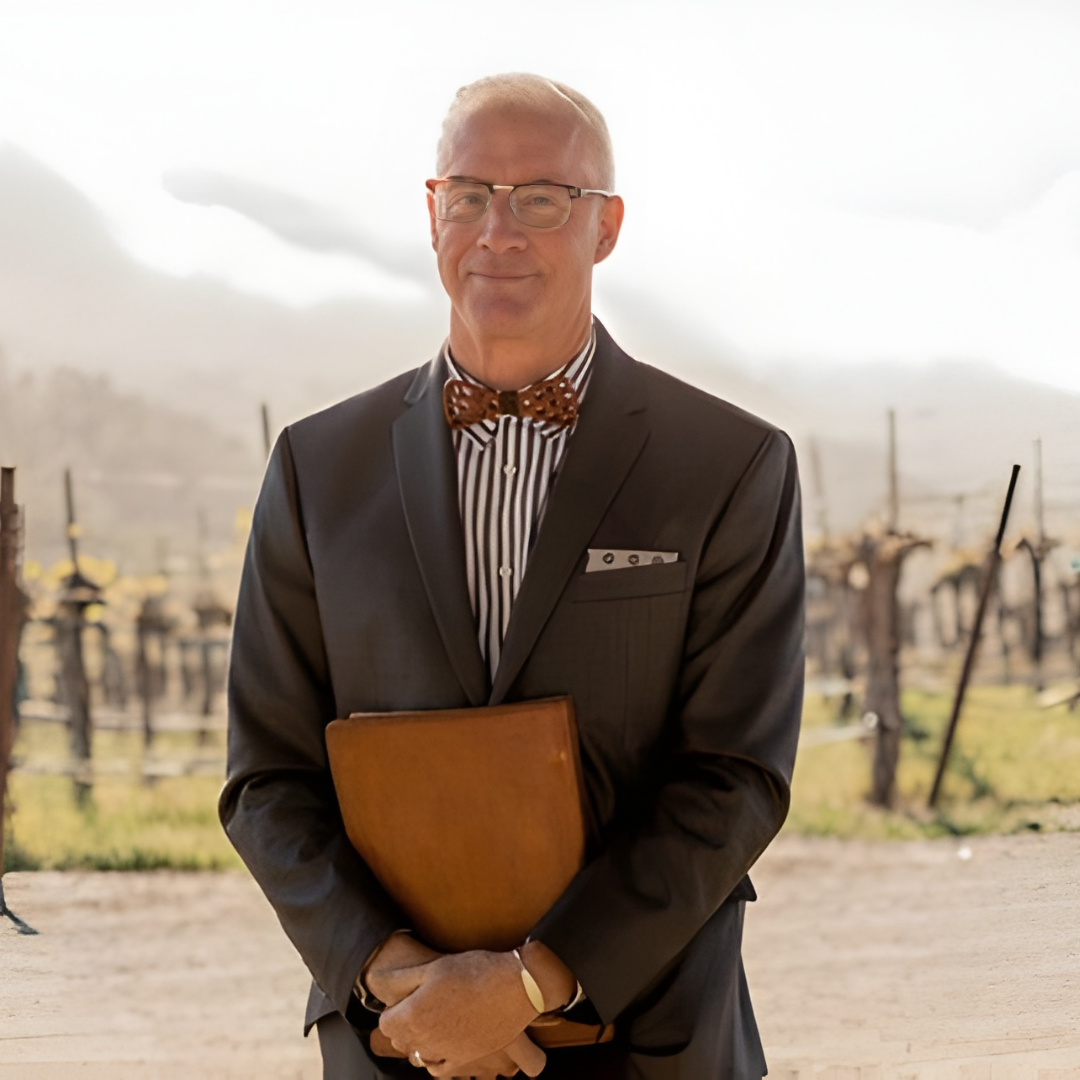 Man in a dark suit and bow tie holding a folder, standing outdoors in bright sunlight