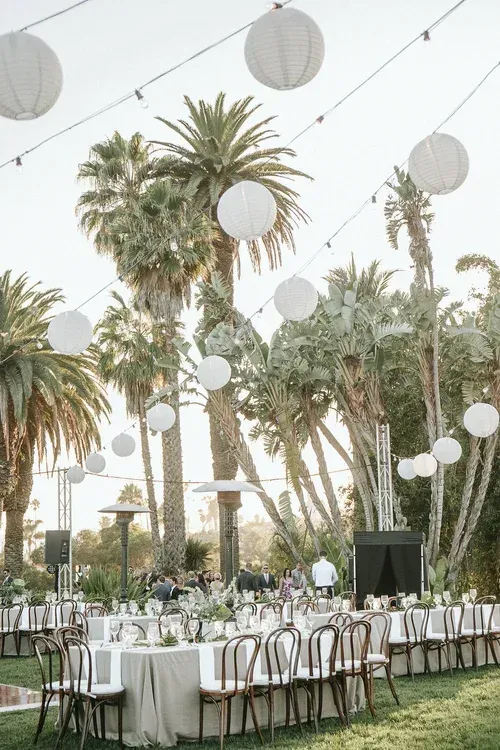Outdoor banquet setup beneath tall palm trees with white lanterns and long tables set for dining.