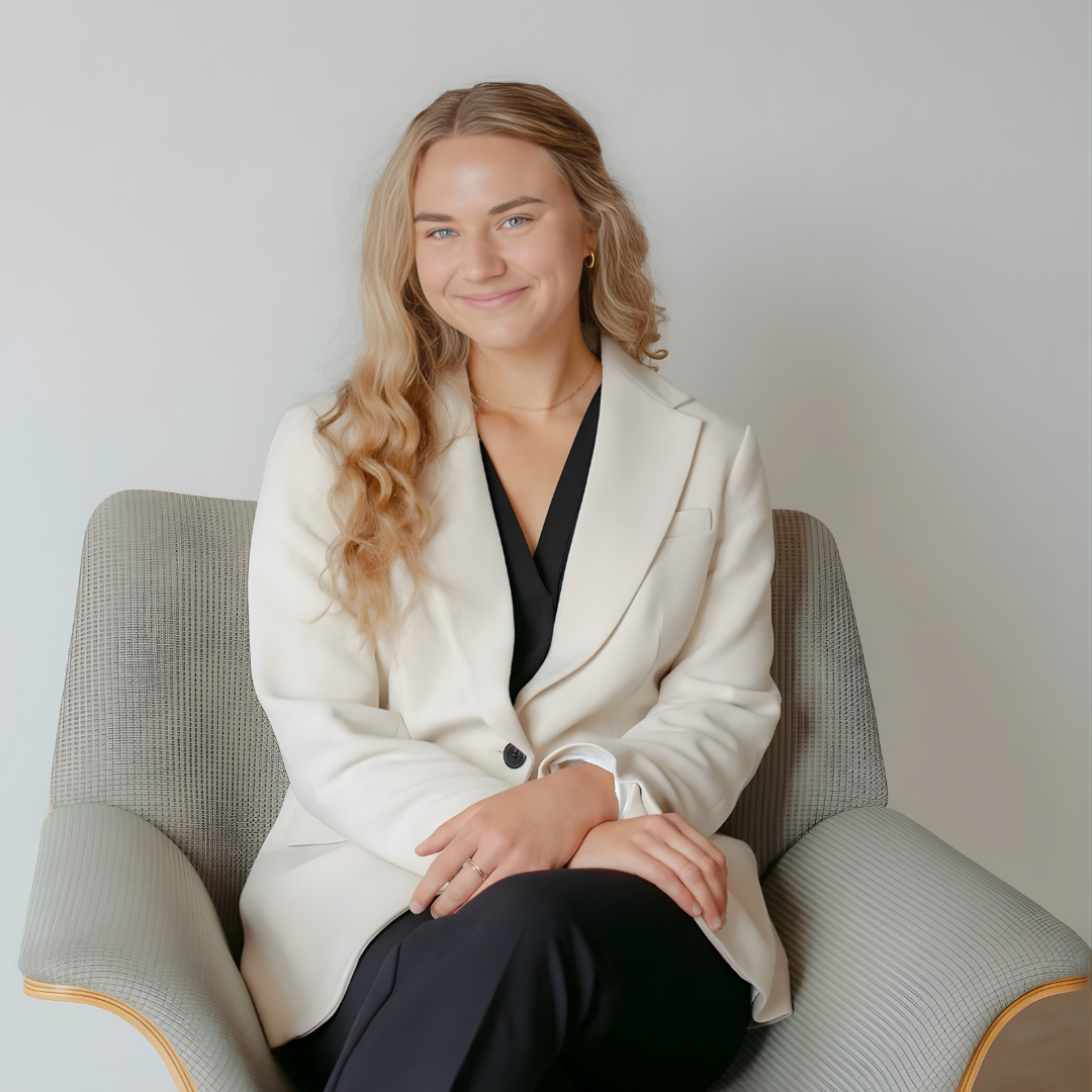 Woman seated in a gray armchair, wearing a white blazer and black dress, smiling gently.