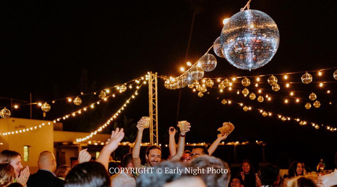 Crowd dancing under string lights at a nighttime outdoor party with a mirrored disco ball overhead