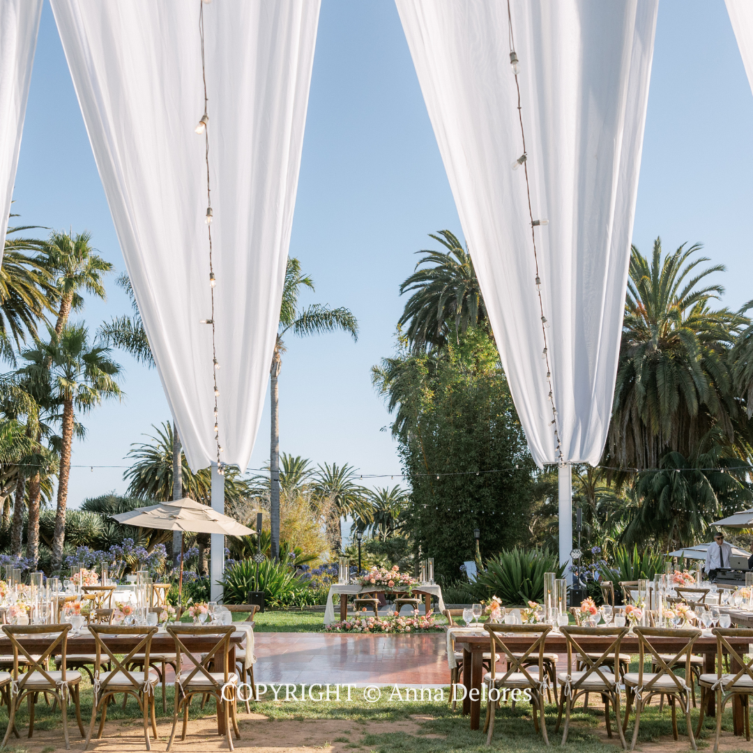 Elegant outdoor dining setup with white drapes, wooden tables, and palm trees around a poolside patio