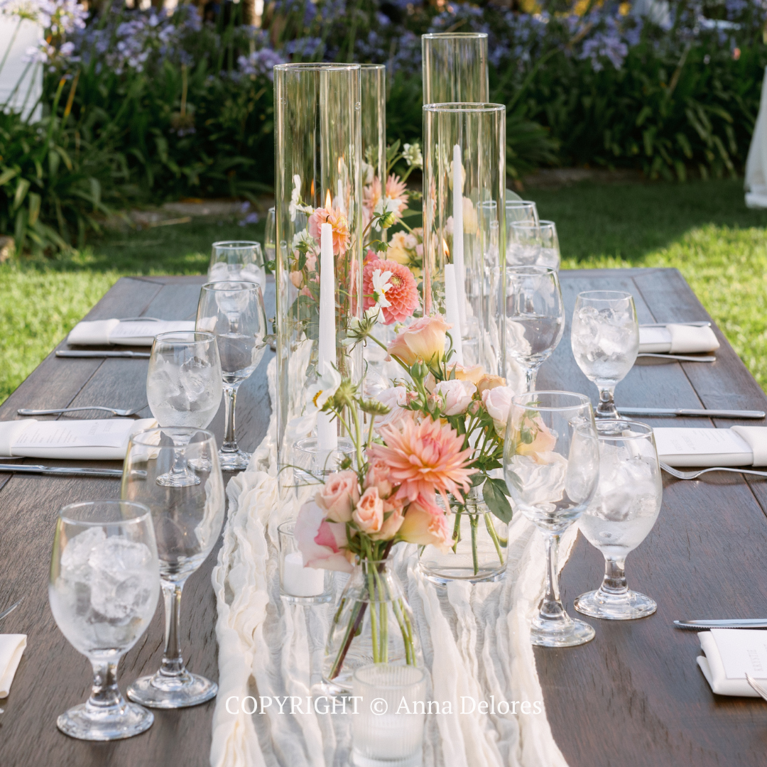 Elegant outdoor dining table with floral centerpiece, tall glass vases, and place settings in a garden.