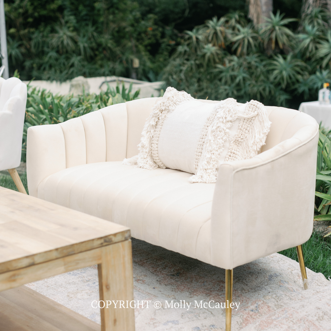 Cream upholstered loveseat with a textured pillow on a patio beside a wooden table and greenery