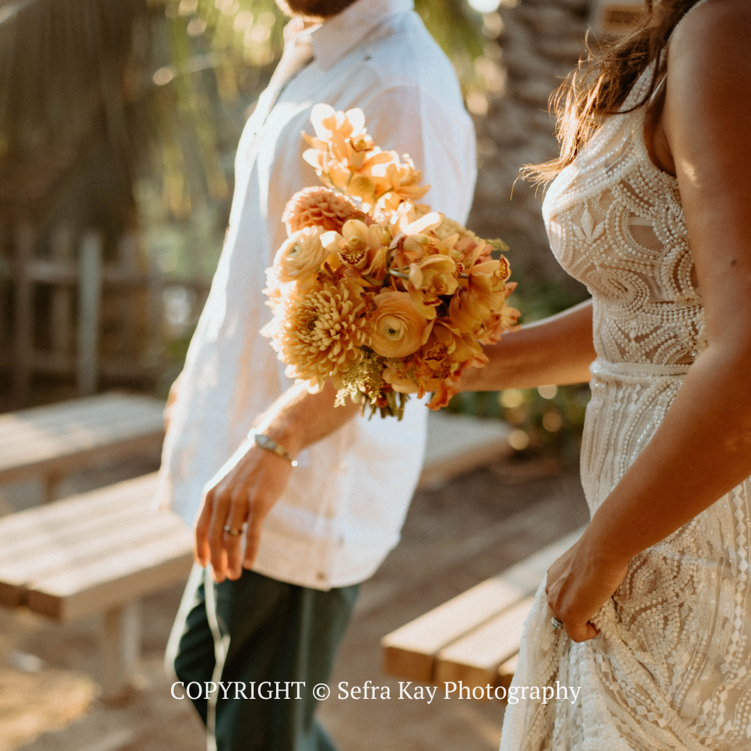 Wedding bouquet held between two women in white dresses outdoors, with warm sunlight.