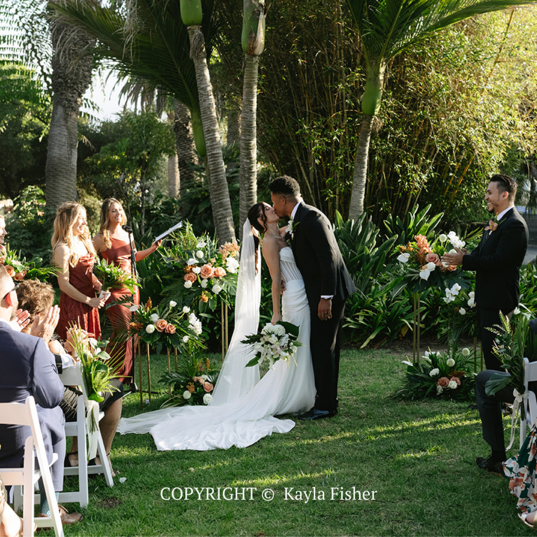 Wedding couple kissing under palm trees in a garden ceremony, with guests seated nearby