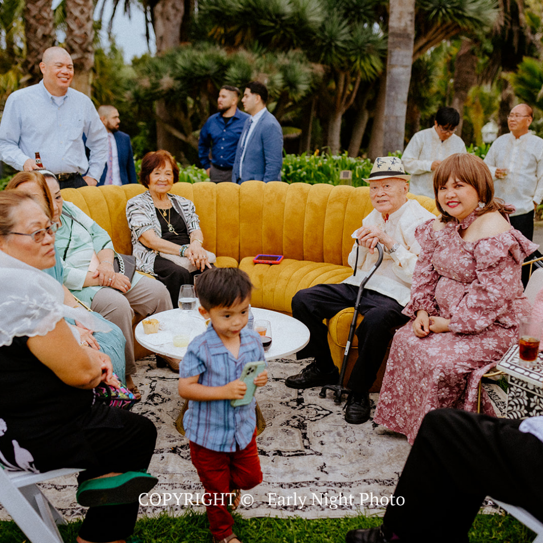 People gathered outdoors on white sofas around a child holding a drink, in a garden setting with yellow seating.