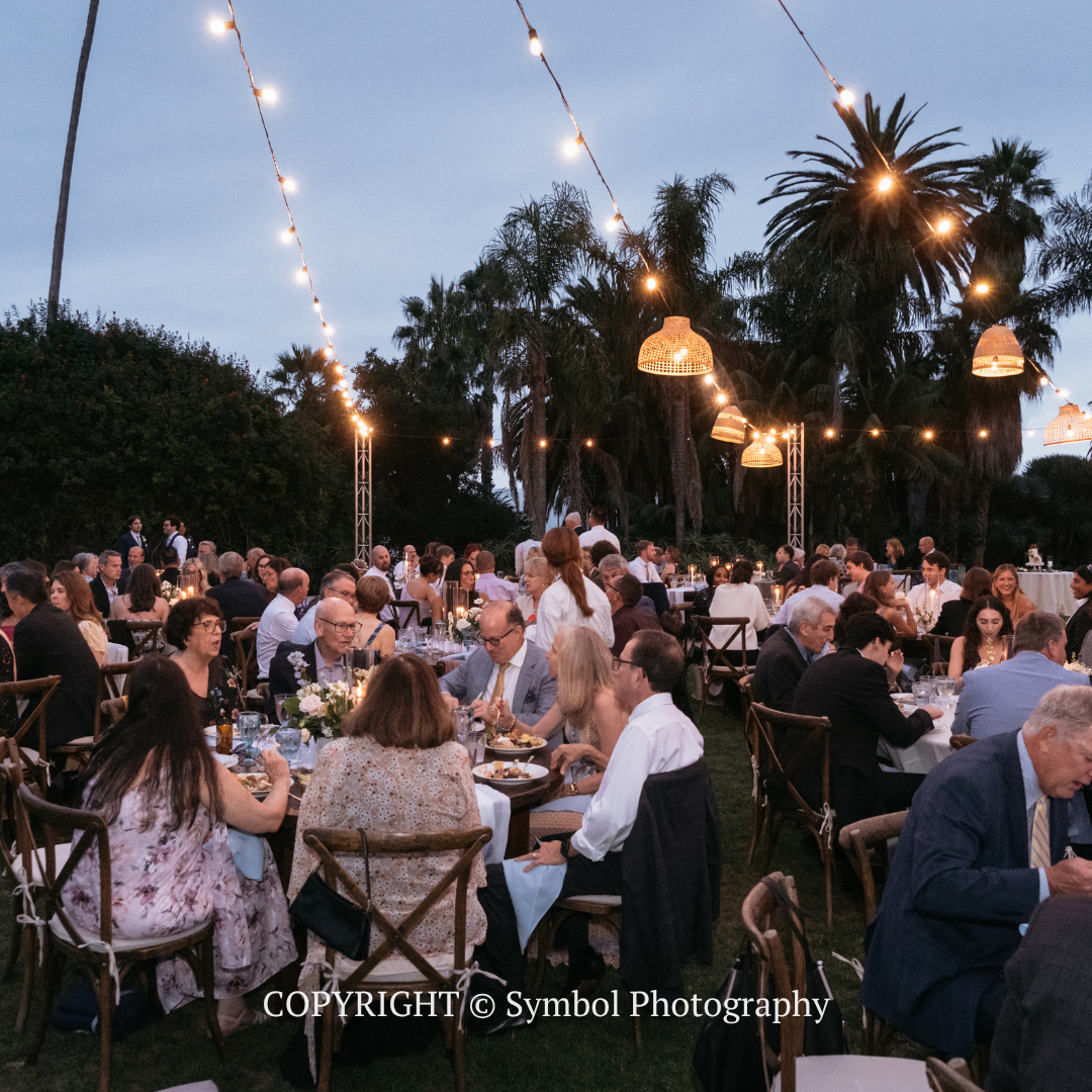Outdoor evening banquet with guests seated at tables under string lights and palm trees
