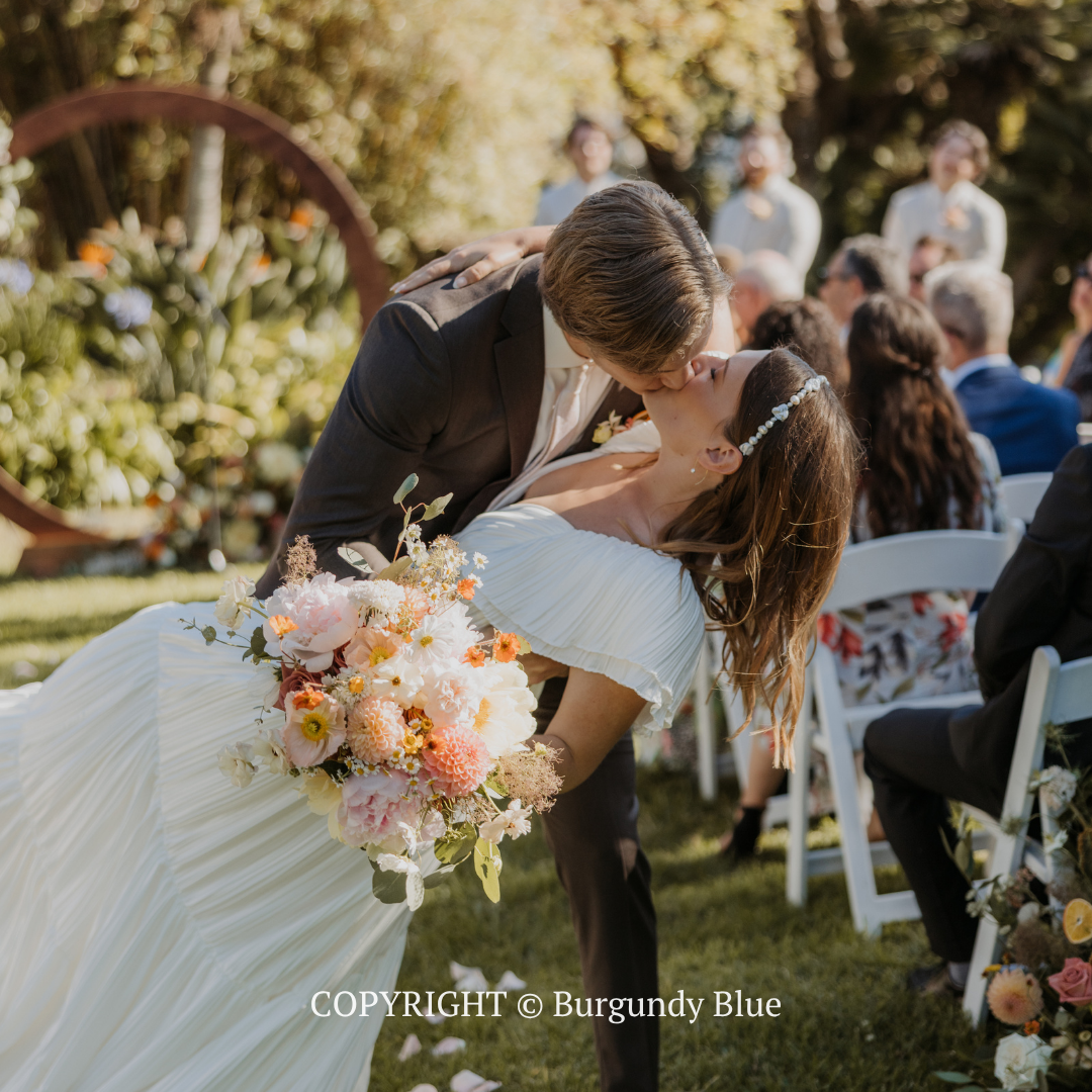 Bride and groom kissing outdoors; bride holds a bouquet beside white chairs and flowers