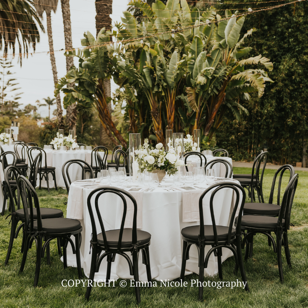 Round outdoor dining tables with white linens and black chairs set in a garden with tall palm trees.