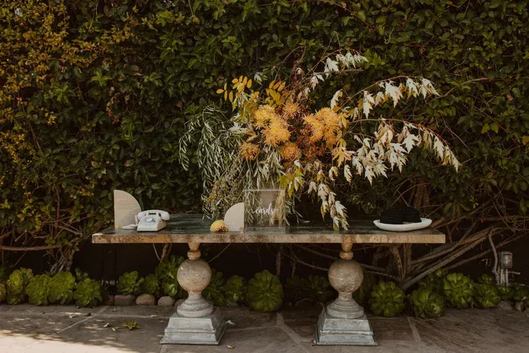 Elegant buffet table with white dishes and tall autumn floral arrangement against a dark leafy wall