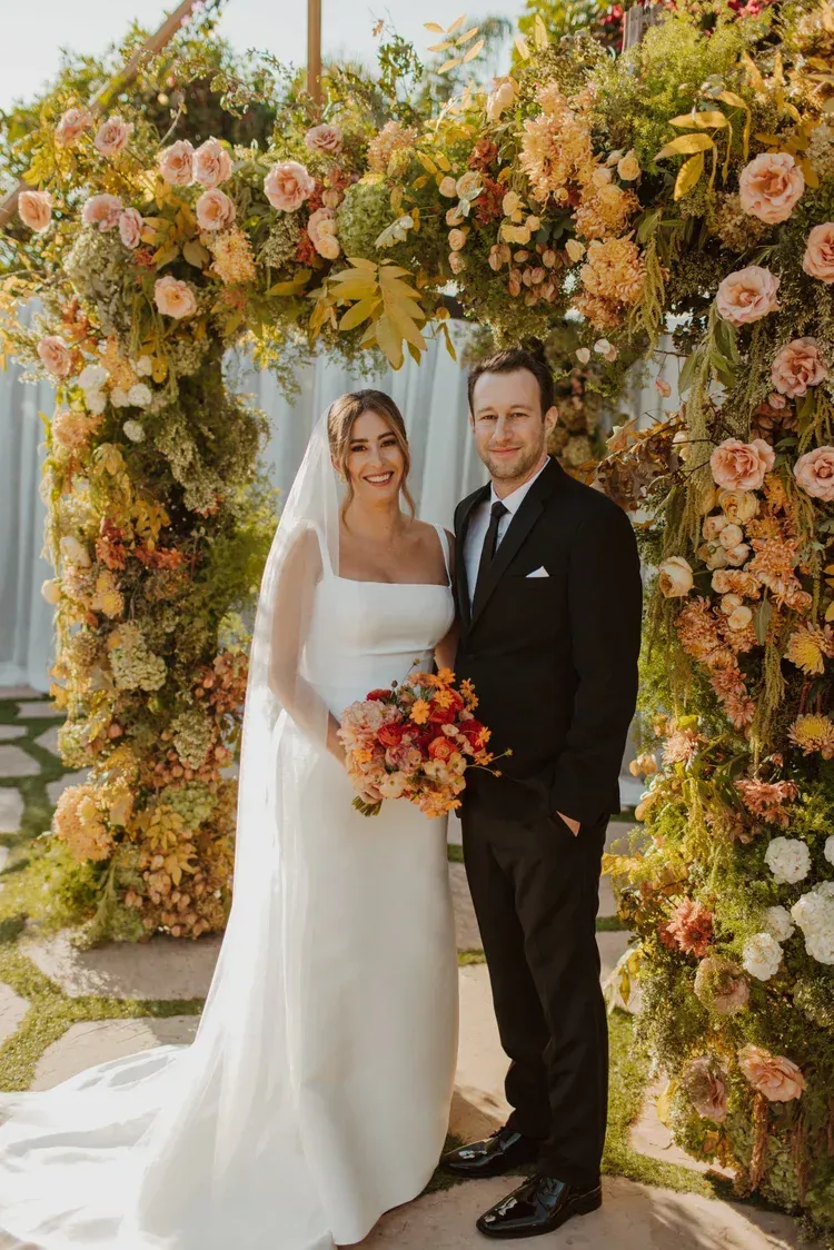 Bride and groom posing under a flower-covered arch, holding a colorful bouquet.