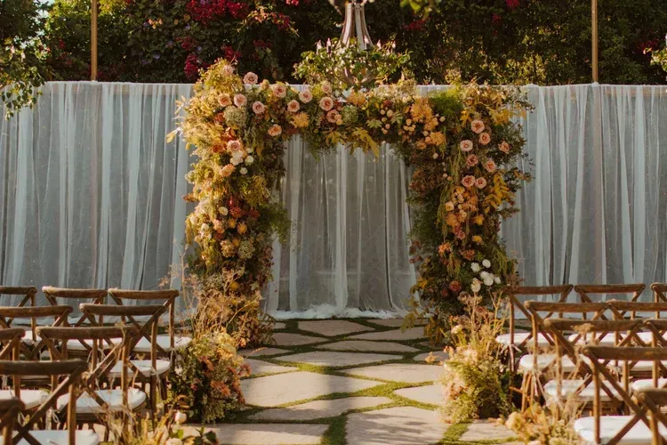 Flower-covered wedding arch lining an outdoor aisle with folding chairs and a draped white backdrop