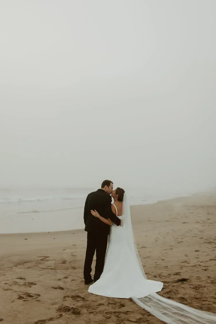 Couple in formal black and white attire embracing on a foggy beach, with a long white dress train on the sand