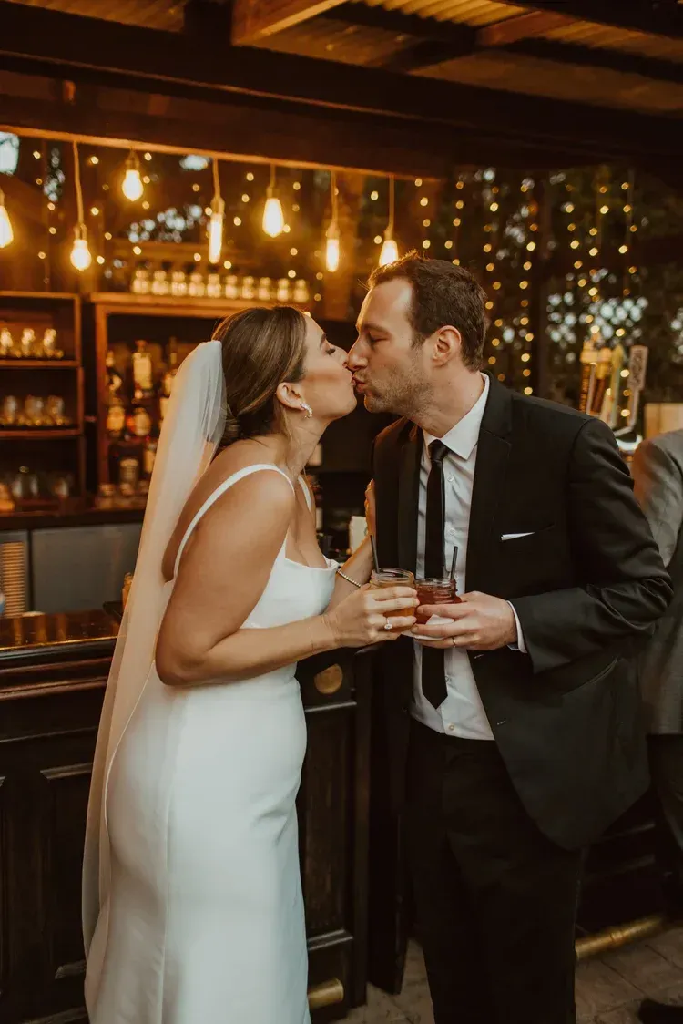 Bride and groom kiss at a warmly lit bar, holding drinks in a festive indoor setting