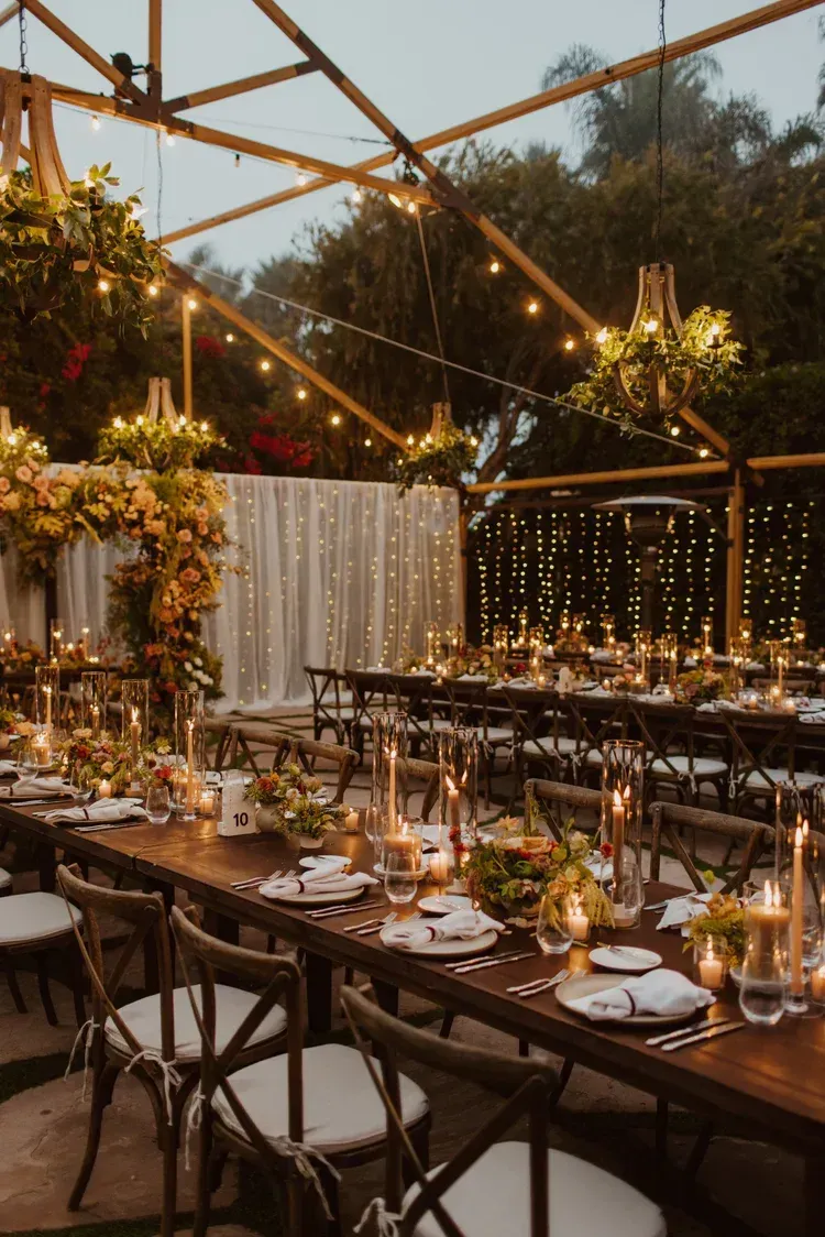 Elegant outdoor dining setup with long wooden tables, candlelight, and floral arrangements under a glass canopy