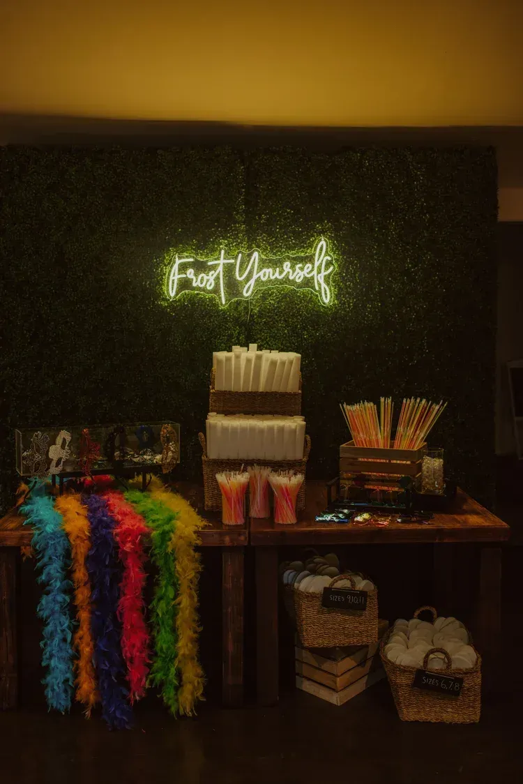Event dessert and favor table with neon sign, rainbow leis, and candy jars against a dark backdrop.