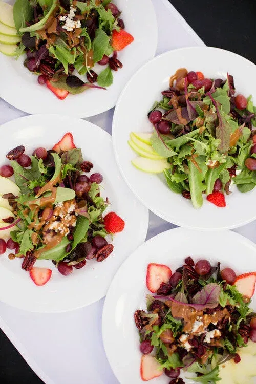 Four plated salads with mixed greens, strawberries, grapes, and lime wedges on a white table