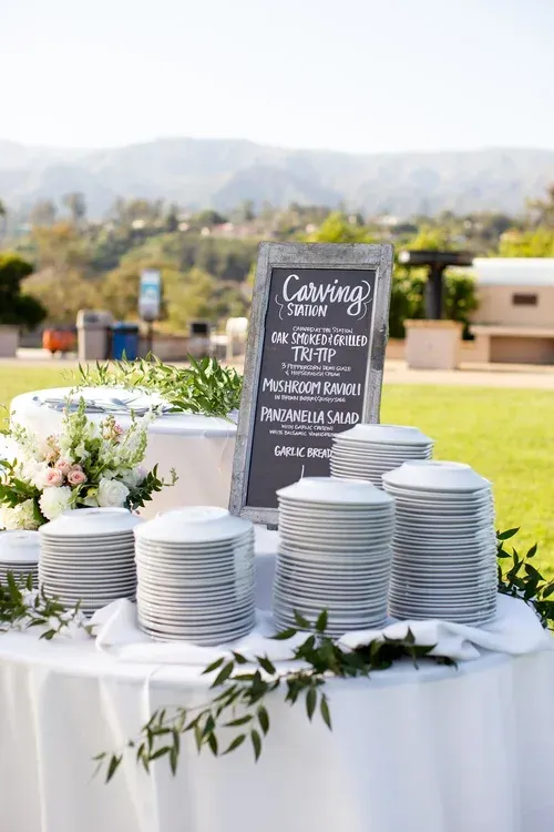 Wedding buffet table with stacked white plates, floral arrangement, and chalkboard sign outdoors