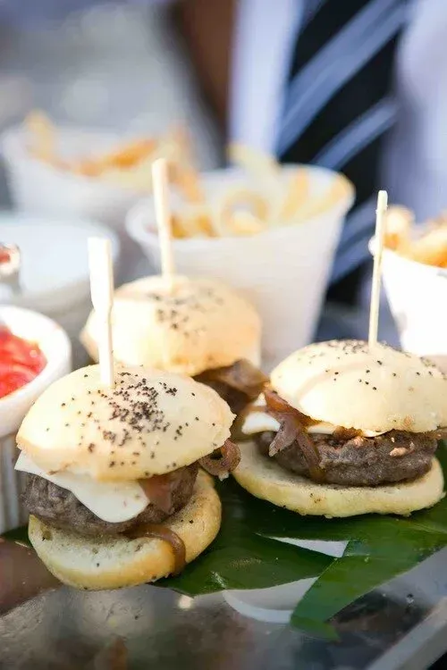 Mini sliders topped with white cheese and sesame buns on a platter with toothpicks and side cups
