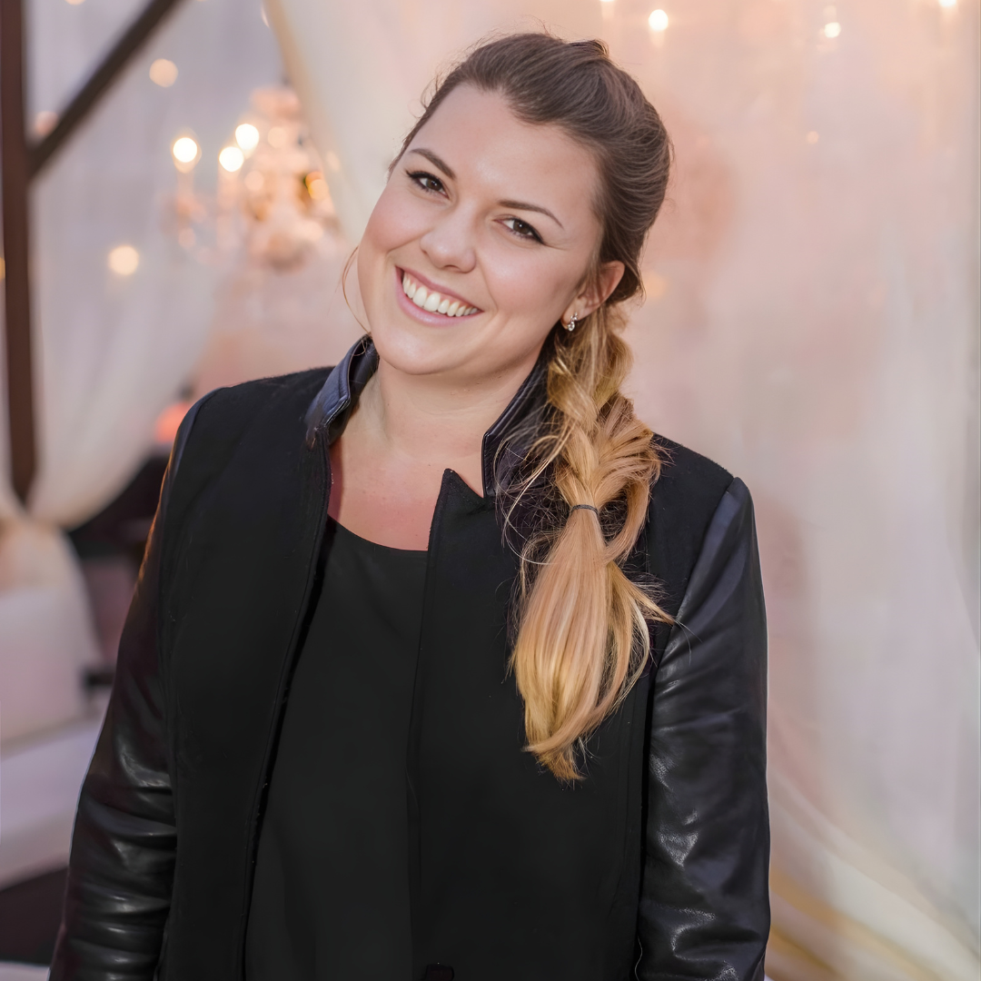 Smiling person in a black jacket with a side braid, standing in a softly lit indoor setting