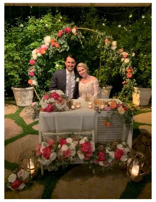 Couple seated at a decorated outdoor wedding table under a floral arch, smiling at the camera.