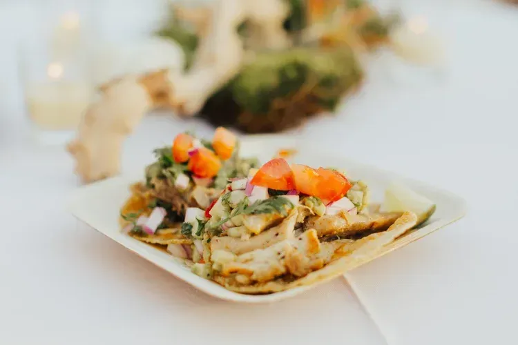 Tacos topped with diced tomatoes and herbs on a white plate at a softly lit table