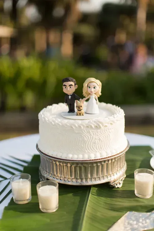Wedding cake with bride and groom figurines on a green tablecloth, surrounded by small candles outdoors