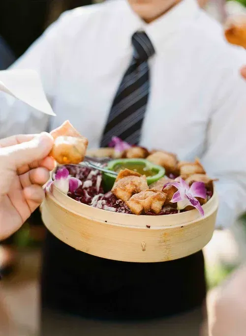 Hands holding a bamboo bowl of fried food and purple garnish, with a blurred waiter in the background