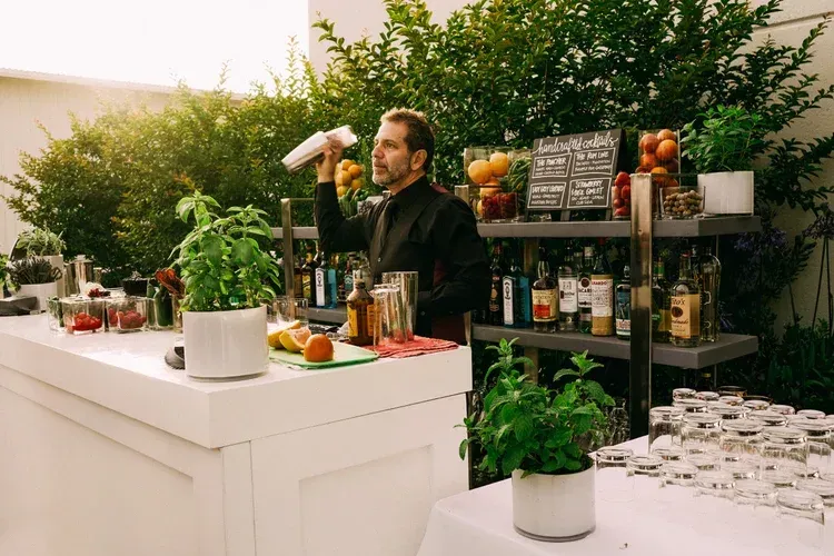 Man behind a white bar counter tasting a drink, with herbs, bottles, and glasses on display.