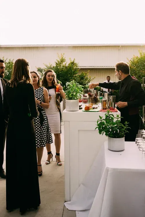 Guests mingling at a cocktail reception beside a white bar with drinks and potted plants