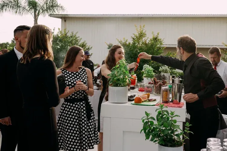 Guests mingling at an outdoor cocktail party around a white bar with drinks and appetizers