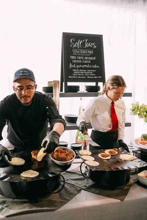 Two chefs cooking at a food stall, stirring pans over a hot griddle with a menu sign behind them